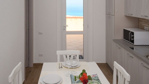 A small, clean room with a dining table set for two, a microwave, and a view of the beach through an open door. - Welcomely - Villa Paola B (Muravera)