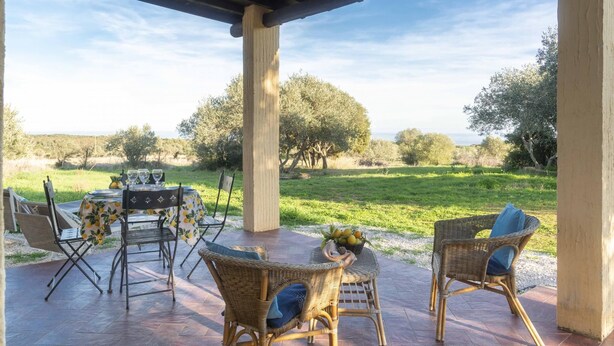 An outdoor patio with wicker furniture, a table set with glasses, and a view of a grassy landscape with trees. - Casa Oliveto 1 in Galtell (Galtelli)
