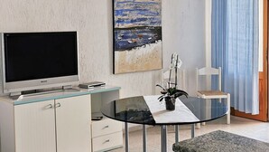 Ein modernes Wohnzimmer mit Flachbildfernseher, einem Gemälde mit Strandmotiv, einem Glastisch mit Blumentopf und einem blauen Vorhang.