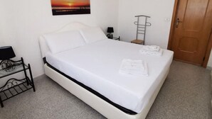 Ein ordentlich bezogenes Bett mit weißen Leinen, ein an der Wand befestigtes Bild eines Sonnenuntergangs, ein Nachttisch mit Lampe und eine Holztür.