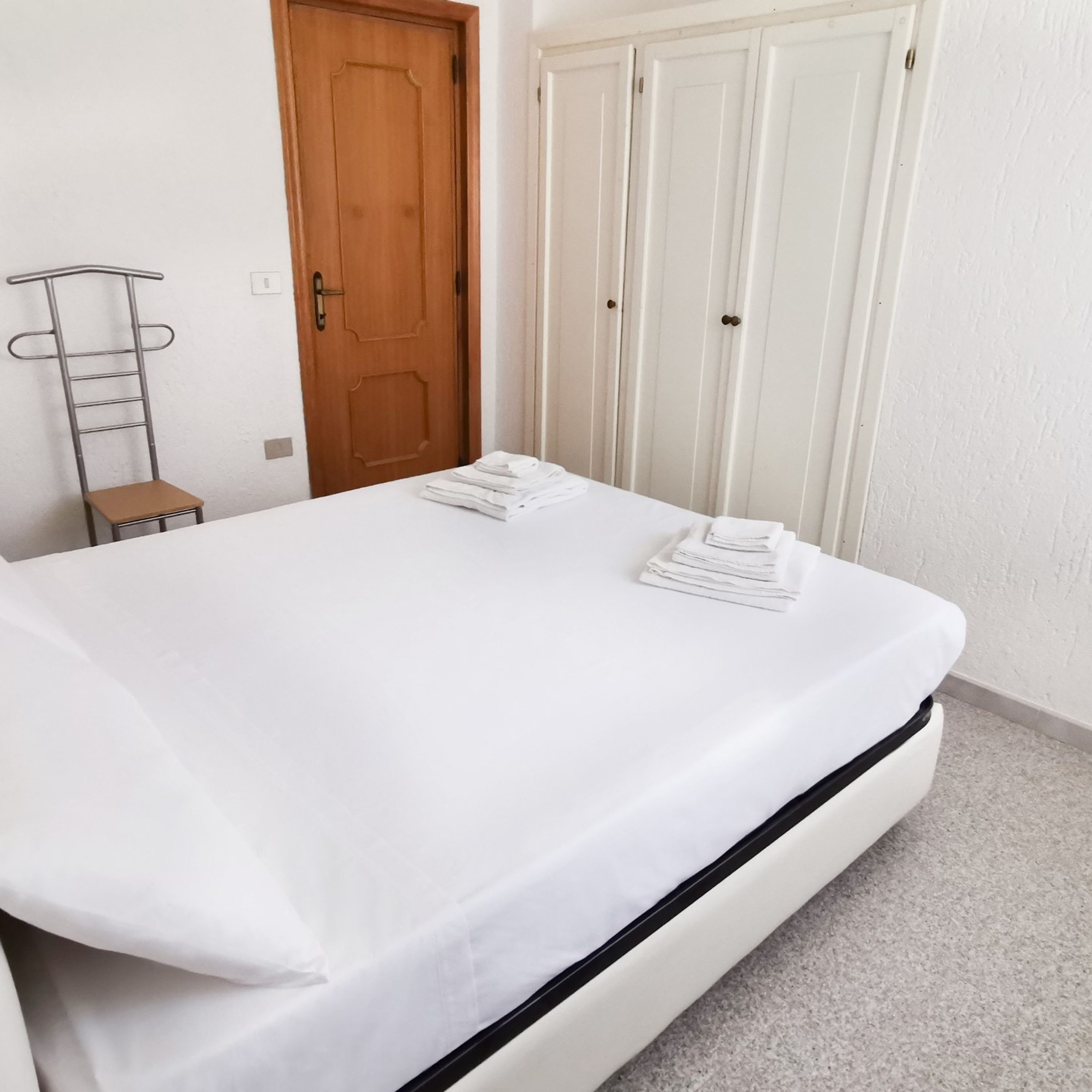 A neatly made bed with white linens, a wooden door, and a metal chair.