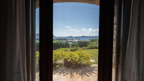 A view through a window with curtains, featuring a garden with yellow flowers, a body of water, and boats. - Welcomely - Villa Acqua Marina (Golfo Aranci)