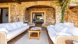 A stone-walled outdoor seating area with two white sofas, a wooden coffee table, and wicker chairs. - Welcomely - Villa Acqua Marina (Golfo Aranci)