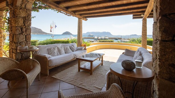 A covered outdoor seating area with stone pillars, wicker furniture, and a view of the sea and boats. - Welcomely - Villa Acqua Marina (Golfo Aranci)