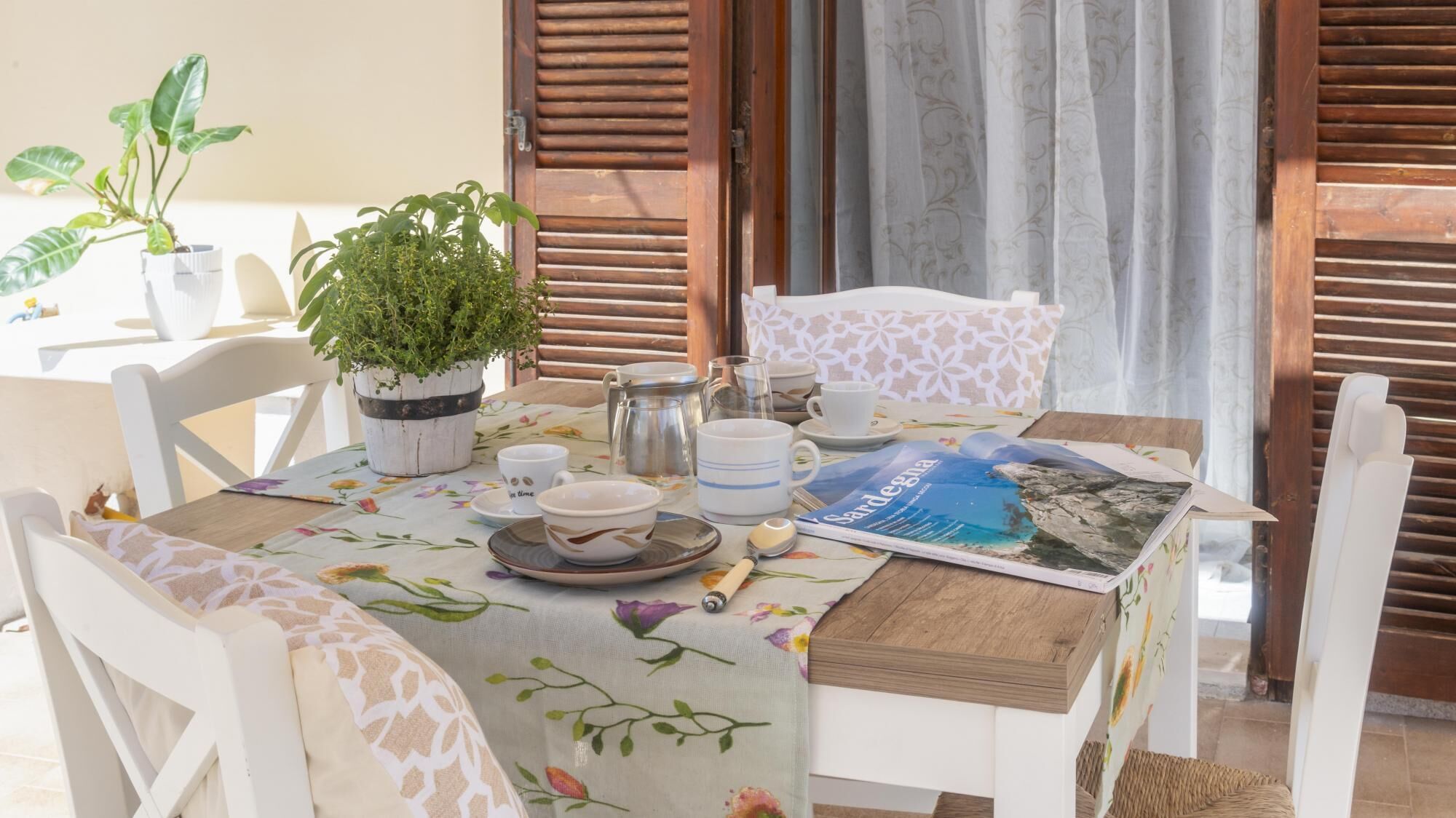 Comedor con una mesa dispuesta para dos personas, mantel con estampado floral, sillas blancas, una planta en maceta y una revista.