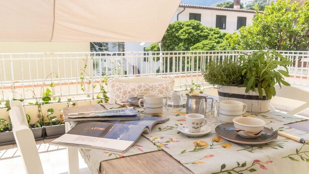 An outdoor dining area with a table set for two, featuring a floral tablecloth, white chairs, and a potted plant. - La Terrazza sul Porto (Dorgali)