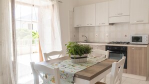 A kitchen with white cabinets, a microwave, a stove, and a sink. There is a dining table with chairs and a potted plant on it. - La Terrazza sul Porto (Dorgali)