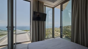A hotel room with a large bed, a flat-screen TV mounted on the wall, and a panoramic view of the coastline through floor-to-ceiling windows. - Porphyra Collection Villas Porto Heli  (Ermionida)