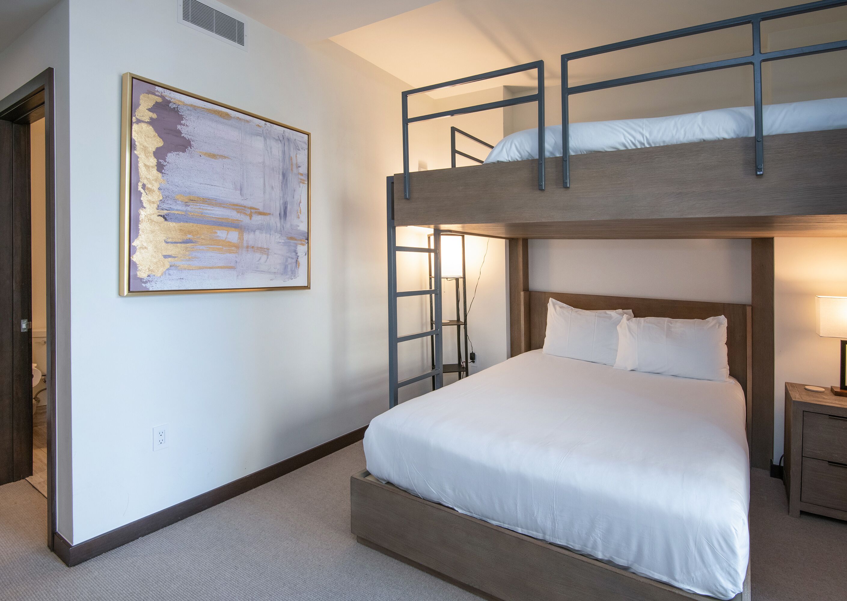 A bunk bed with a wooden headboard and white bedding, a ladder, a bedside table with a lamp, and a painting on the wall.
