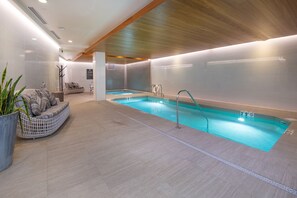 Un espace piscine intérieur moderne, doté d’une piscine à eau claire, de poutres en bois au plafond et d’un coin salon avec des coussins.