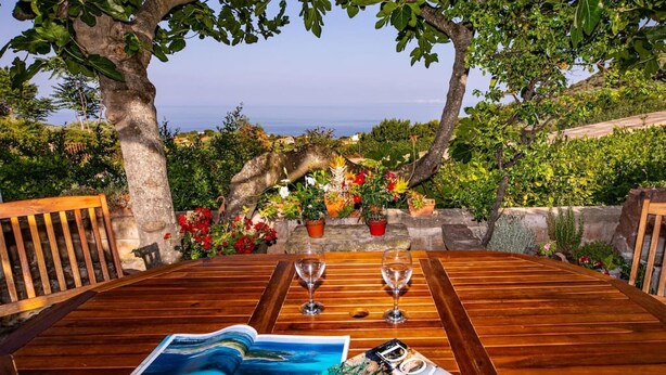 A wooden table with two wine glasses, a magazine, and a view of the sea through a tree-lined terrace. - Welcomely - Badde Nuraghe (Magomadas)