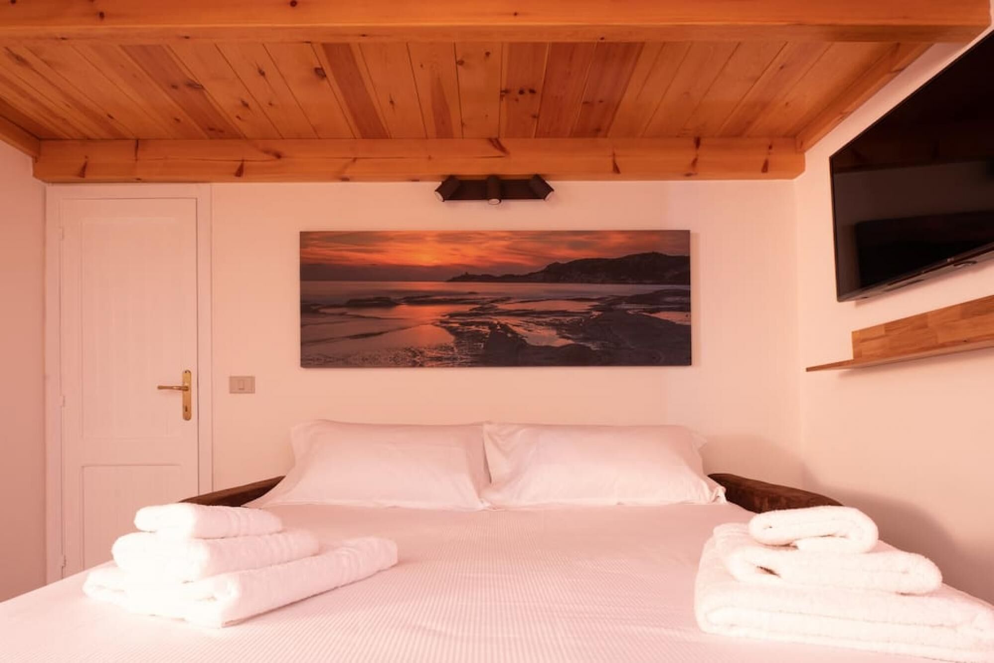 Quarto com teto de madeira, cama com roupa de cama branca, pintura de uma paisagem ao pôr do sol e uma TV fixada na parede.