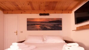 A bedroom with a wooden ceiling, a bed with white bedding, a painting of a sunset landscape, and a TV mounted on the wall. - Welcomely - Badde Nuraghe (Magomadas)