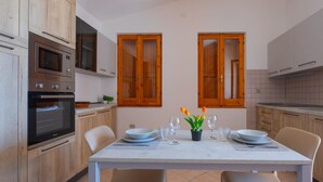 A modern kitchen with wooden cabinets, a built-in oven, and a dining table set for two with chairs. - Welcomely - Badde Nuraghe (Magomadas)