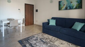 A living room with a dark blue sofa, two teal cushions, a white dining table with chairs, a wooden door, and a painting of fish on the wall. - Welcomley - Xenia Boutique House - Apt 5 (Olbia)