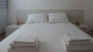 A neatly made bed with white linens, two bedside tables with lamps, and a window with blinds. - Welcomley - Xenia Boutique House - Apt 5 (Olbia)
