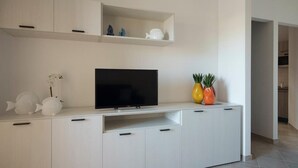 A modern living room with a flat-screen TV on a white entertainment unit, a small shelf with decorative items, and a doorway leading to another room. - Welcomley - Xenia Boutique House - Apt 5 (Olbia)