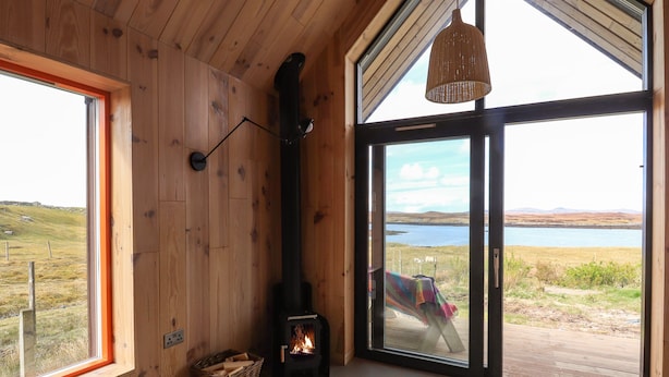 A cozy room with wooden walls, a large window, a fireplace, and a view of a lake. - North Light Studio (Isle of North Uist)