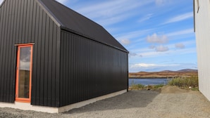 A black building with a red door, located next to a body of water and hills in the background. - North Light Studio (Isle of North Uist)