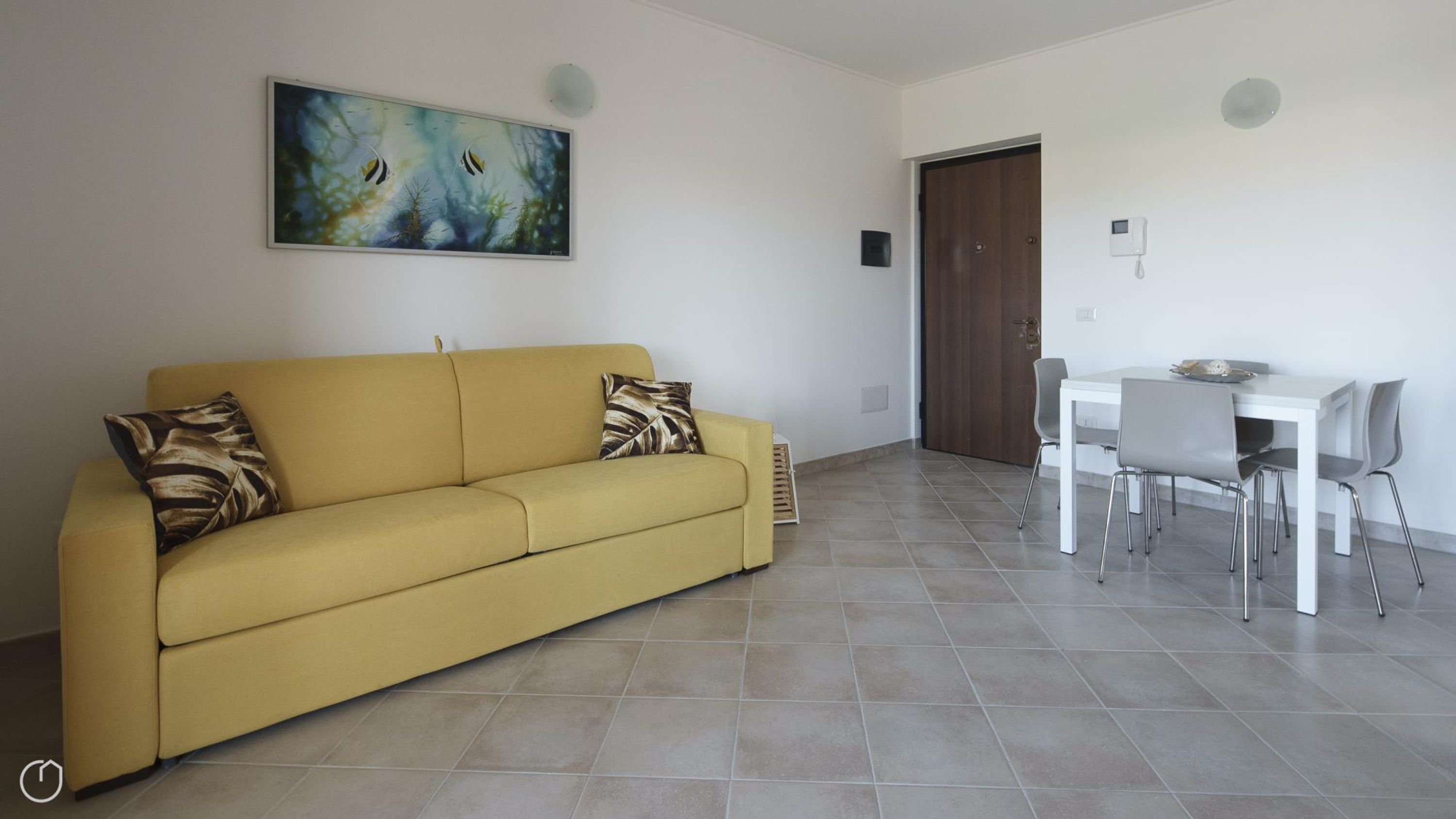 A yellow sofa with patterned cushions, a dining table with chairs, and a painting on the wall.