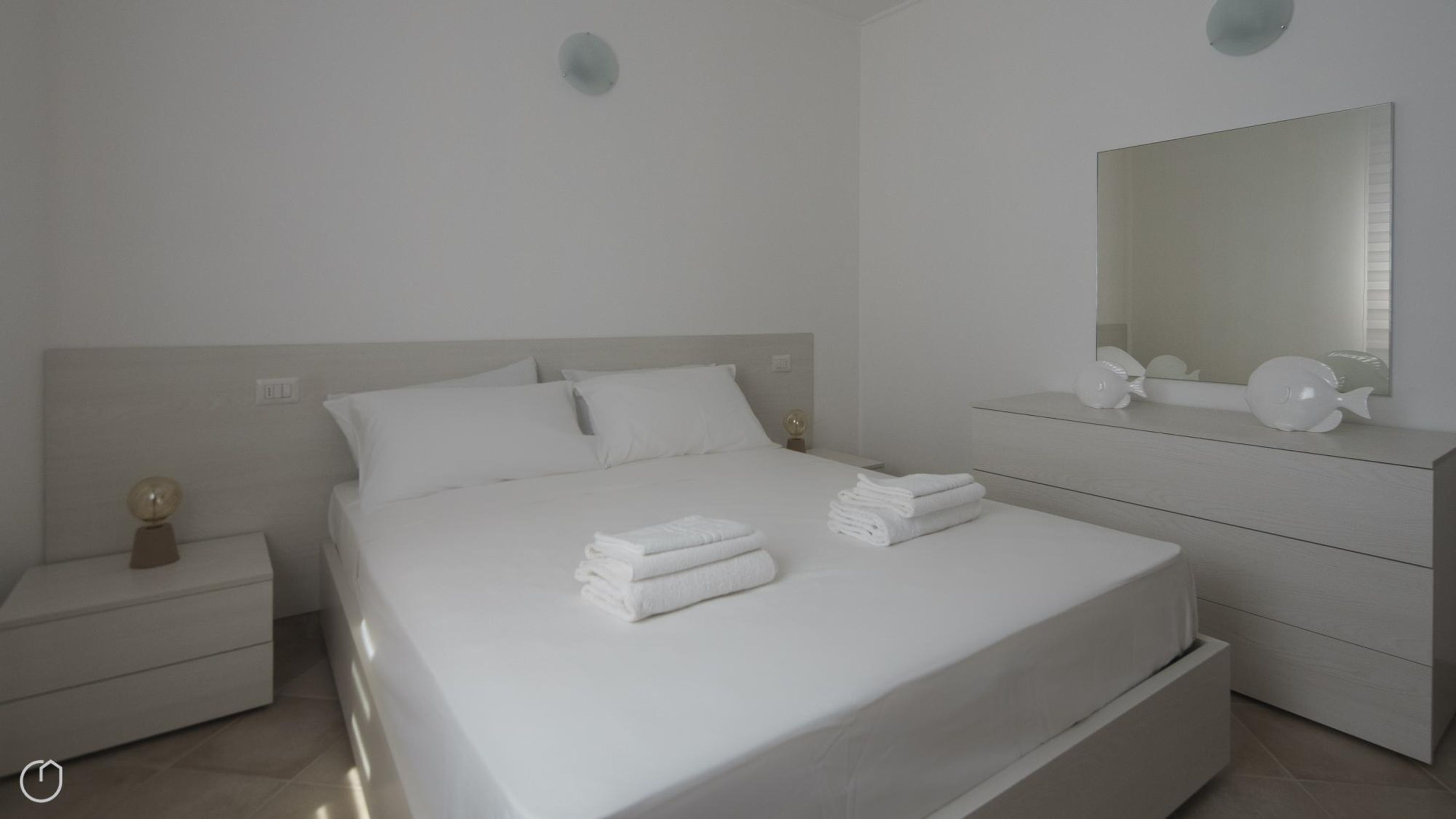 A modern, minimalist bedroom with a large bed, a bedside table with a globe, a dresser with a mirror, and a wall-mounted light fixture.