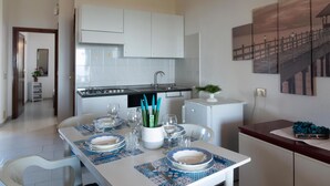 A modern kitchen with white cabinets, a stove, and a refrigerator. There is a dining table set with plates, glasses, and a vase with green plants. - Welcomely - Casa Della Luna (Budoni)