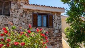 A stone building with a tiled roof, wooden shutters, and a window with patterned curtains. - Welcomely - Casa Della Luna (Budoni)