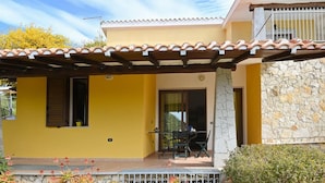 A yellow building with a tiled roof, a stone wall, and a patio area with a table and chairs. - Welcomely - Villa Alba (Muravera)