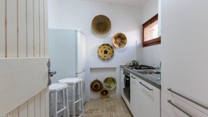 A compact kitchen with white cabinets, a gas stove, a sink, and a refrigerator. There are stools and decorative plates on the wall. - Welcomely - Porto Cervo sul Mare (Arzachena)