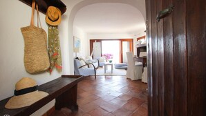 A cozy living room with a bench, a hat, and a woven bag on the wall. A wooden door leads to a dining area with a table and chairs. - Welcomely - Porto Cervo sul Mare (Arzachena)