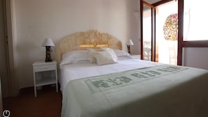 A neatly made bed with a decorative headboard, two bedside tables with lamps, and a view of a balcony with flowers. - Welcomely - Porto Cervo sul Mare (Arzachena)