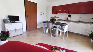 Uma cozinha moderna com armários brancos, uma mesa de jantar e duas cadeiras. Há uma televisão num móvel, uma planta e um sofá vermelho em primeiro plano.