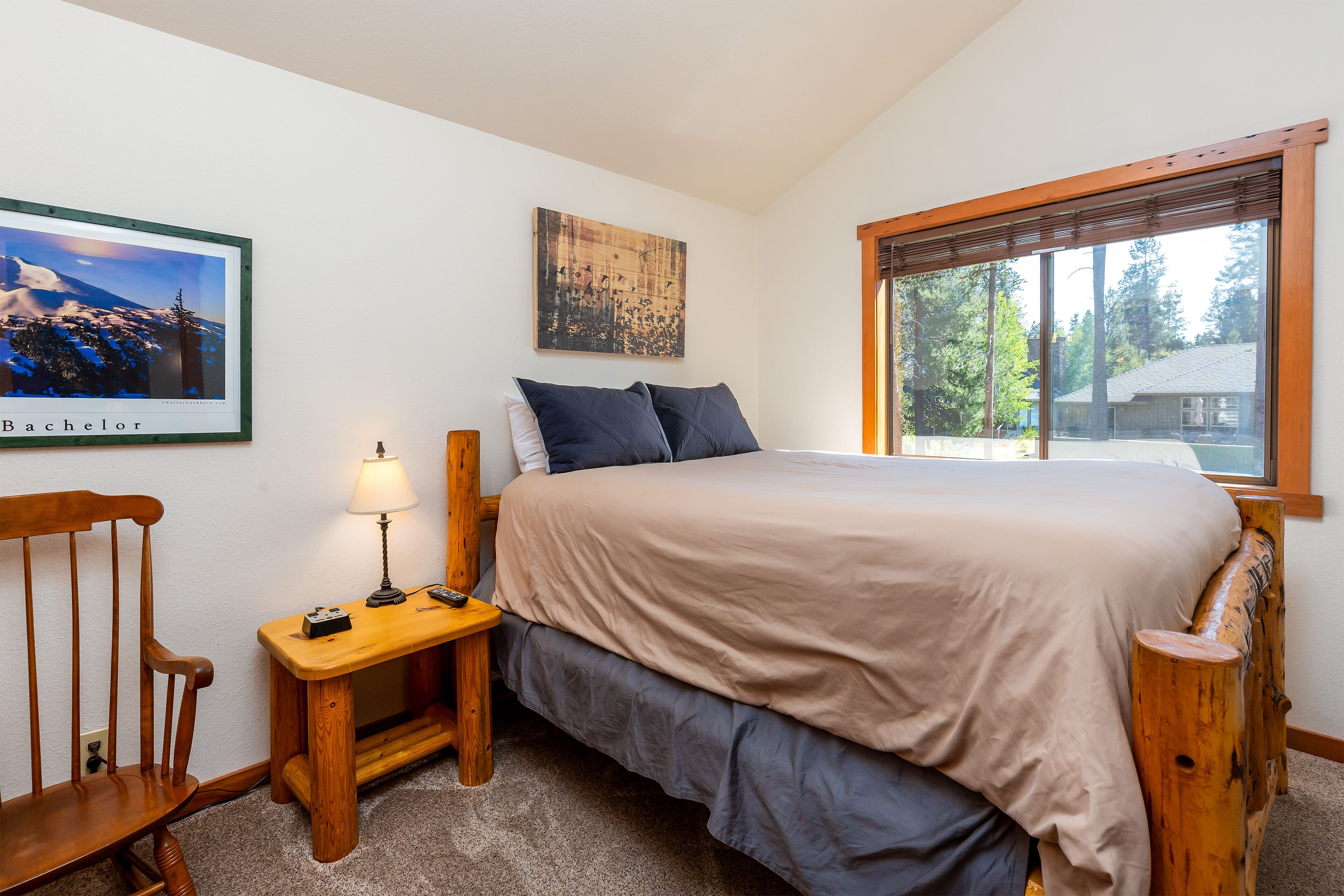 A bedroom with a bed, a nightstand, a chair, a lamp, and a window with a view of trees.