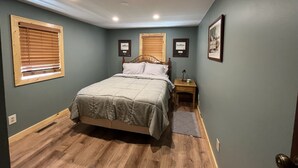 A bedroom with a bed, two bedside tables, a nightstand, a window with blinds, and a framed picture on the wall.