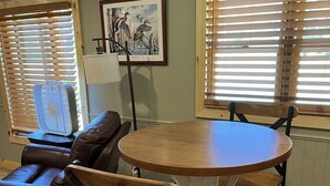 A dining area with a round wooden table, four chairs, a brown leather sofa, and two windows with blinds.