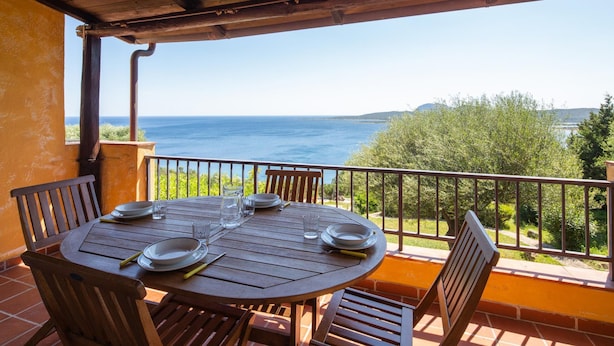 Ein runder Holztisch mit vier Stühlen, gedeckt mit Tellern, Gläsern und Besteck. Der Tisch steht auf einem Balkon mit Blick auf das Meer und Grünflächen.