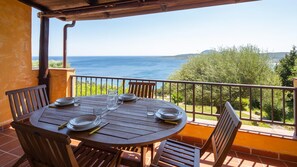 A round wooden table with four chairs, set for dining with plates, glasses, and cutlery. The table is on a balcony with a view of the sea and greenery. - Welcomely - Porto Rotondo sul Mare (Olbia)
