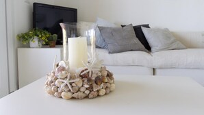 Ein weißer Tisch mit einem Glas-Kerzenhalter, der eine Kerze und Muscheln enthält, steht auf einer weißen Tischdecke. Im Hintergrund ist eine weiße Couch mit grauen Kissen und ein gerahmtes Bild an der Wand zu sehen.