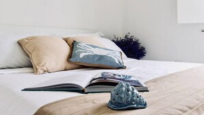 Ein ordentlich gemachtes Bett mit einer beigen Tagesdecke, zwei Kissen, einem offenen Buch und einem Paar Schuhen.