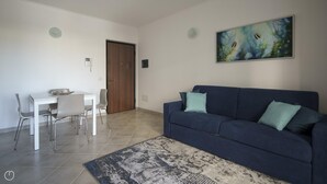 A living room with a dark blue sofa, a white dining table with chairs, and a painting on the wall. - Welcomely - Xenia Boutique House - Apt 7 (Olbia)