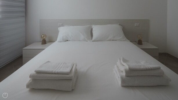 A neatly made bed with white linens, two bedside tables with lamps, and a wall-mounted light switch. - Welcomely - Xenia Boutique House - Apt 7 (Olbia)