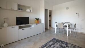 A modern kitchen with white cabinetry, a flat-screen TV, and decorative items. There is a dining area with a white table and chairs. - Welcomely - Xenia Boutique House - Apt 7 (Olbia)