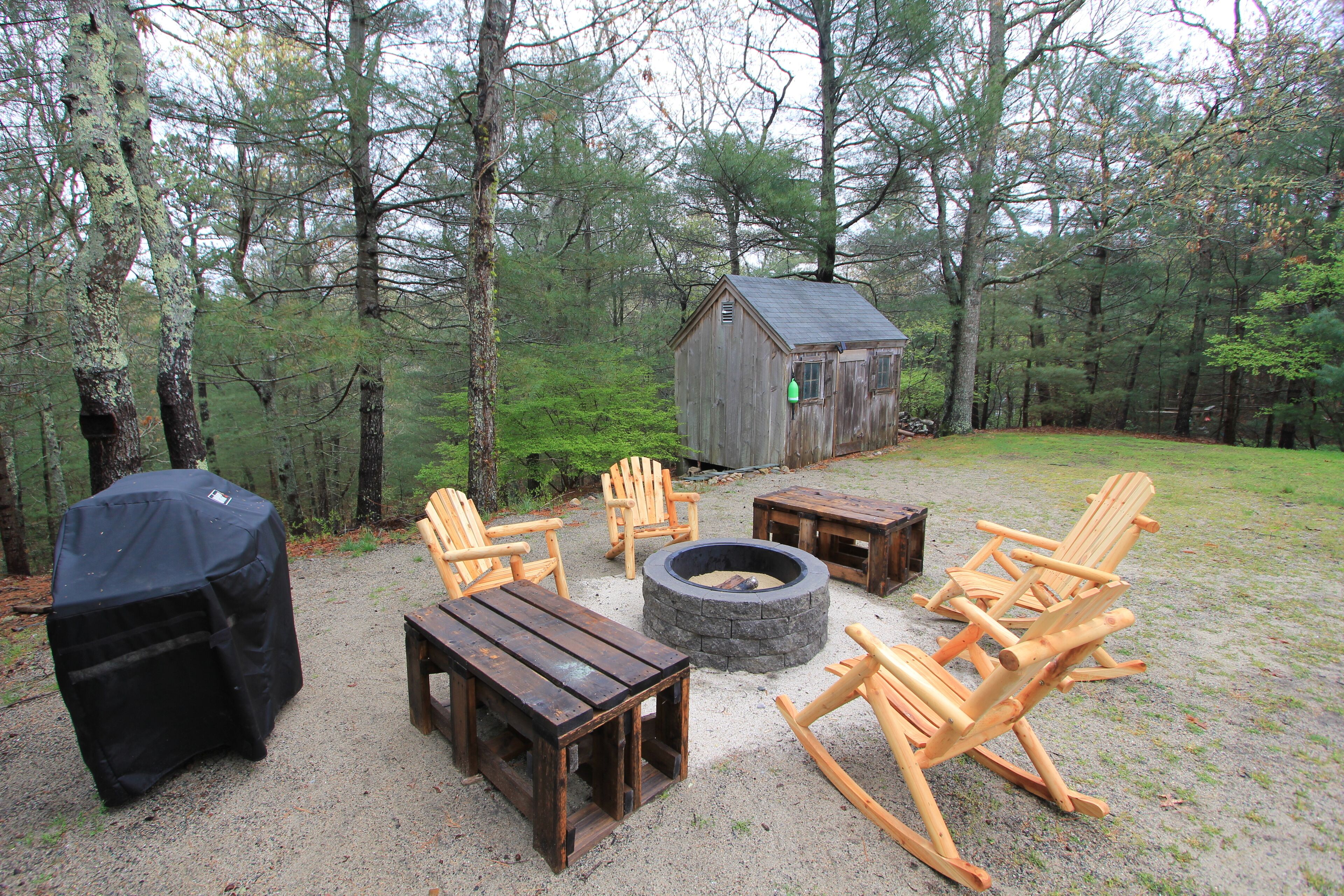 A rustic outdoor seating area with wooden furniture, a fire pit, and a small wooden structure in a forest setting.