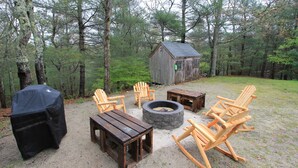A rustic outdoor seating area with wooden furniture, a fire pit, and a small wooden structure in a forest setting.