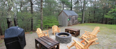A rustic outdoor seating area with wooden furniture, a fire pit, and a small wooden structure in a forest setting.