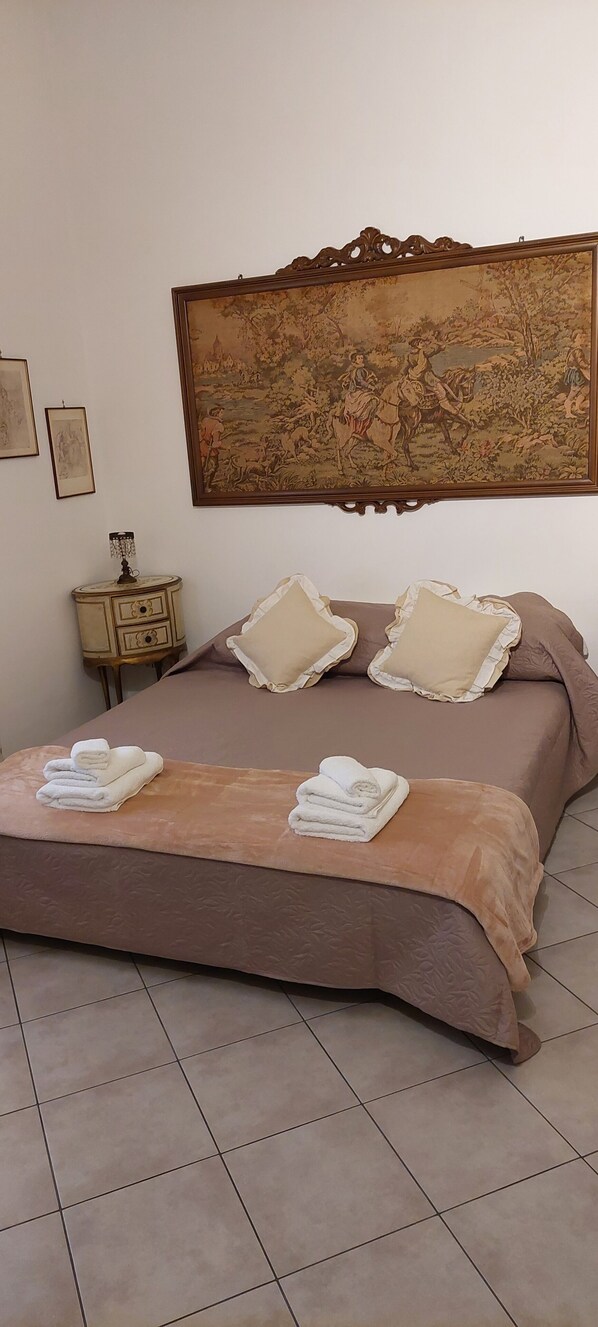A neatly made bed with a brown bedspread and two pillows, a small bedside table with a lamp, and a large tapestry on the wall.