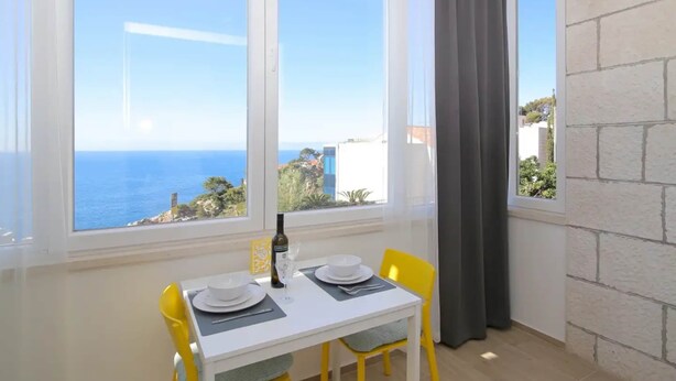 A dining area with a table set for two, a bottle of wine, and a glass. The room has a large window with a view of the sea and buildings. - Apartment Miriam - Studio Apartment With Sea View (Dubrovnik)