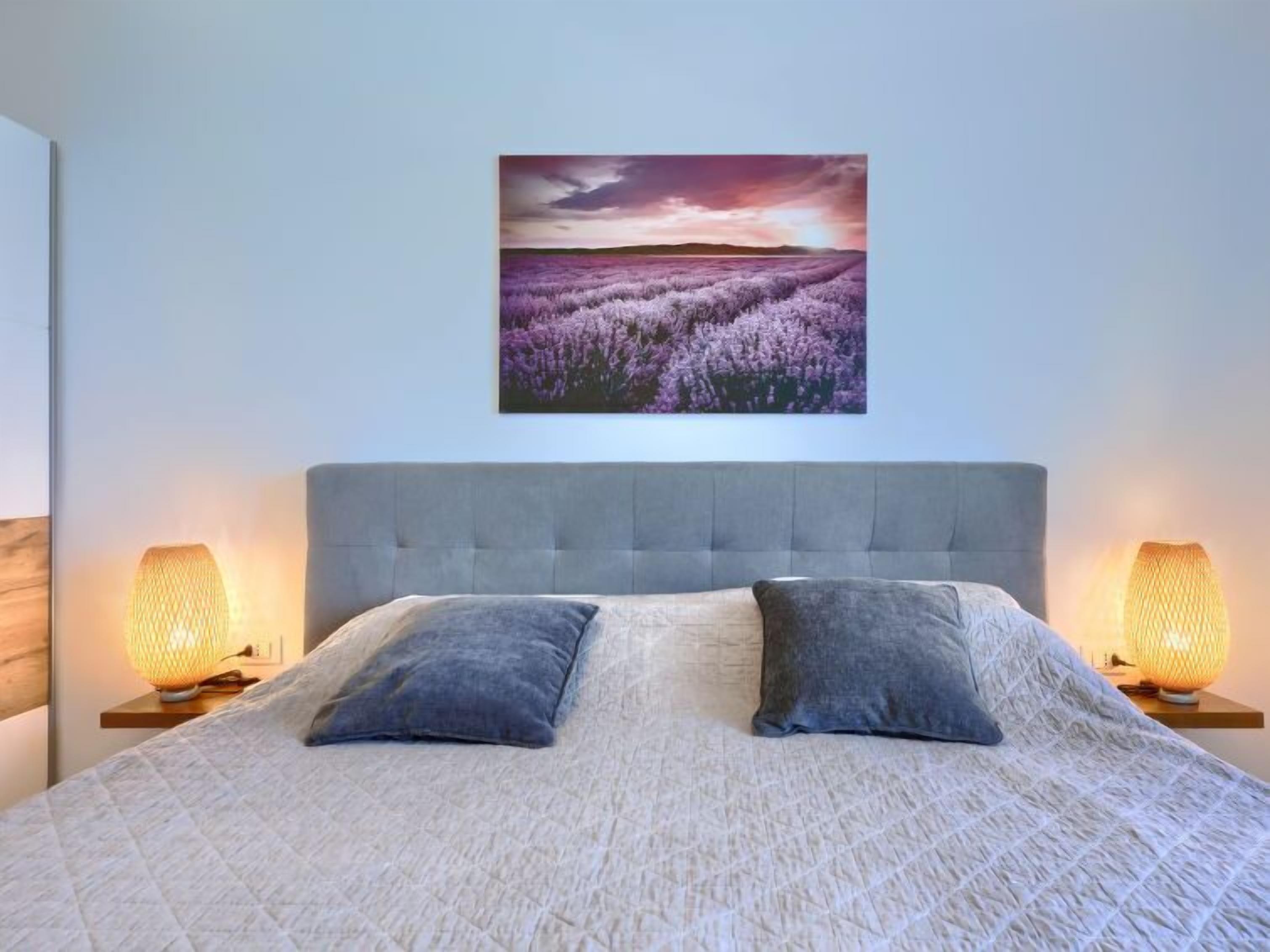 A bedroom with a large bed, two bedside lamps, and a wall-mounted picture of a lavender field.