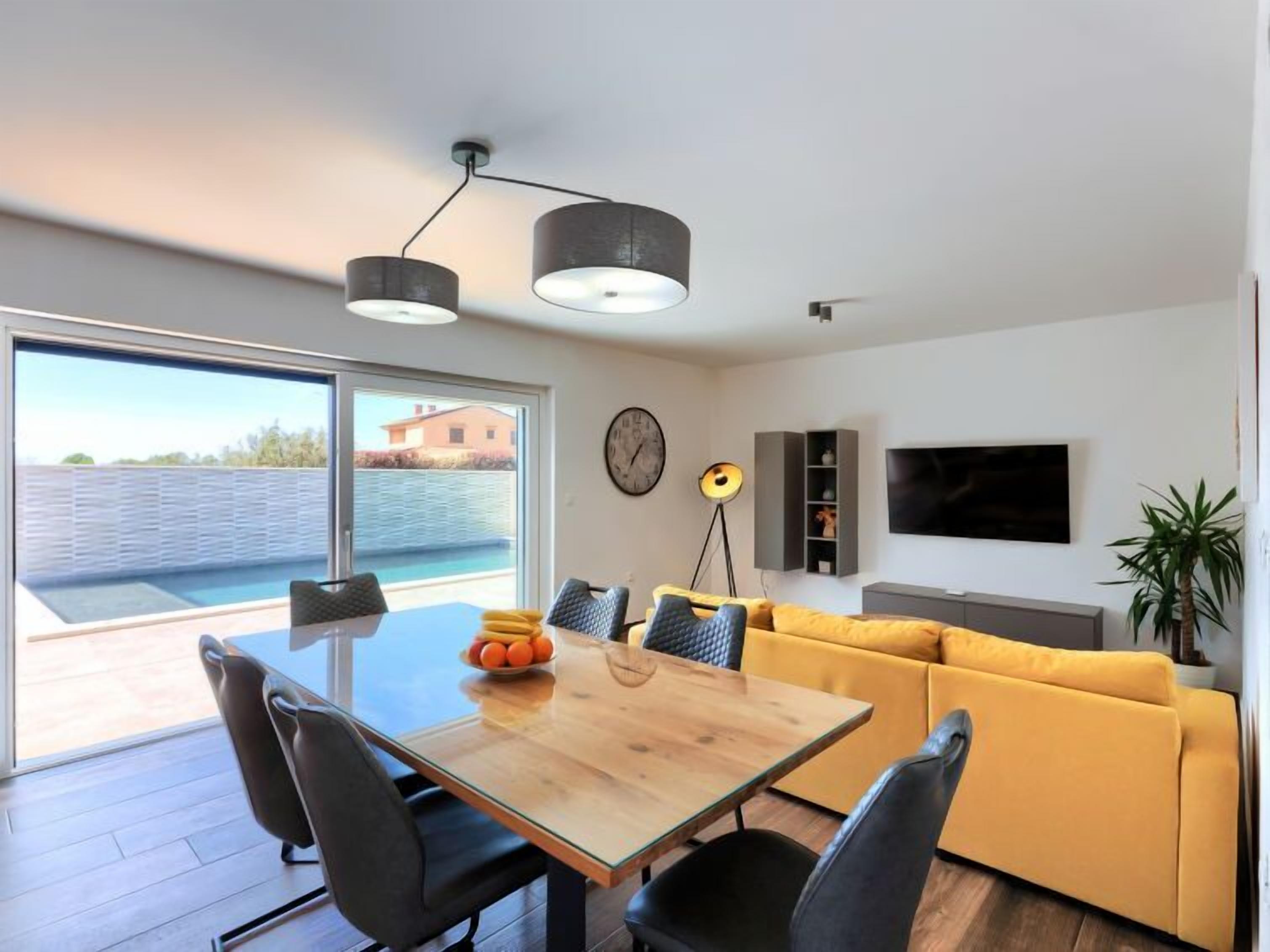A modern living room with a wooden dining table, black chairs, a yellow sofa, and a TV mounted on the wall.