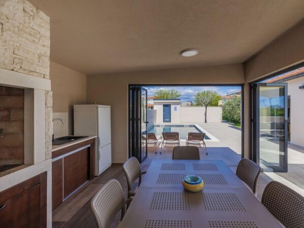 A compact kitchen with a built-in oven and a sink, a dining area with a table and chairs, and a view of a swimming pool and a building in the background.